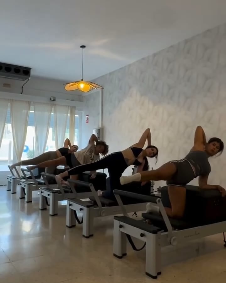 Clase de reformer en la sala de Los Cristianos: cinco alumnas trabajan de lado sobre las camas, junto al ventanal con cortinas de lino y bajo la lámpara en forma de flor